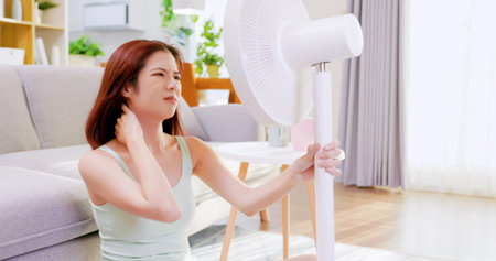 asian unhappy woman is sitting in the front of working fan suffering from summer heat at home