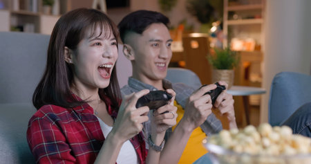 excited Asian young gamer couple sitting on couch holding wireless controller playing video games on...