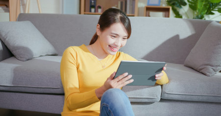 Young asian woman using digital tablet to watch movie happily while sitting on floor when relax at...