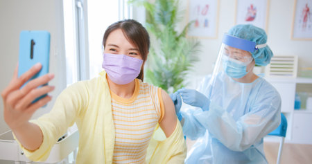 Smiling Asian woman wearing face mask taking selfie with mobile phone while receiving coronavirus...