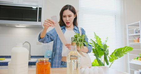 inflation concept - worried asian woman is looking at her shopping receipt from her cotton Eco bag and shocked about the price at kitchenの写真素材