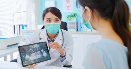 asian woman wearing protective face mask to prevent COVID19 has uterus utltrasonographic diagnosis in hospital - female doctor showing test results to patient on digital tabletの写真素材