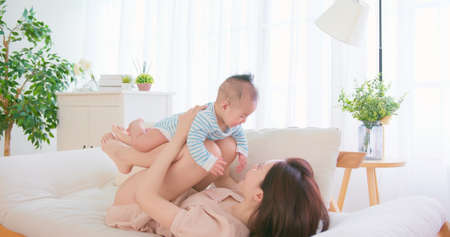 authentic shot of asian mother plays with her baby happily on sofa by lifting infant up at homeの写真素材
