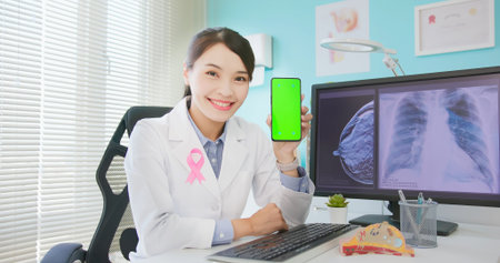 asian female doctor wearing pink ribbon with mammography test results and xray on computer monitor in hospital - she holds green screen mobile phoneの写真素材