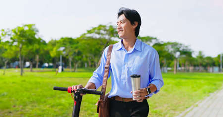 asian businessman walks with electric scooter and holds reusable eco-friendly ecological cup while commuting in the cityの写真素材