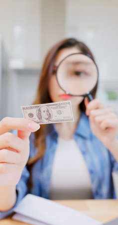 inflation concept - worried asian woman find money is getting smaller by magnifier at kitchenの写真素材