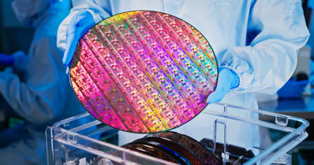 close up of asian male technician in sterile coverall holds wafer with gloves that reflects many different colors and check it at semiconductor manufacturing plantの写真素材