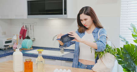 inflation concept - worried asian woman is looking at empty wallet and shocked with her cotton Eco bag at kitchenの写真素材