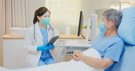 Senior people healthcare concept - asian female doctor with digital tablet is talking to senior man patient lying on bed in hospitalの写真素材