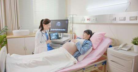 asian pregnant woman lying in hospital ward has uterus utltrasonographic diagnosis - female doctor shows picture on computer and digital tabletの写真素材