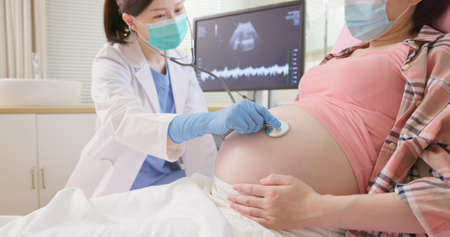 asian female doctor exams a pregnant woman lying in hospital ward by stethoscope - they are wearing protective face mask to prevent COVID19の写真素材