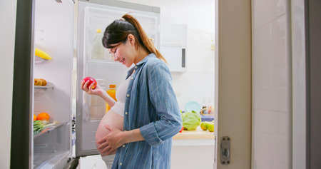 Proper nutrition concept - asian pregnant woman is opening refrigerator and pick out apple in kitchen at homeの写真素材