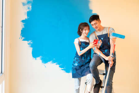 young asian couple decorate home - they take selfie by mobile phone and looking photos while painting wall with blue colorの写真素材