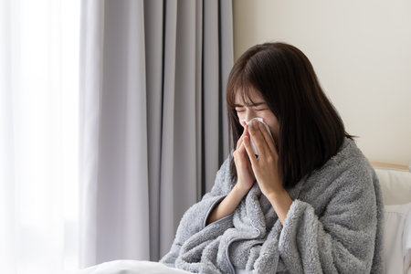 sick asian woman is blowing running nose and sneezing in tissue on bed at homeの写真素材