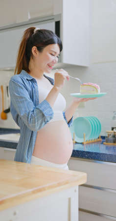 unhealthy eating concept - Asian pregnant woman eats cake in kitchen at homeの写真素材