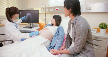 asian pregnant woman lying on bed has ultrasound scan with husband and doctor explains the results in hospitalの写真素材