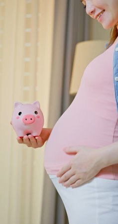 close up of asian pregnant woman holding a piggy bank standing near window at home - Saving for Baby...