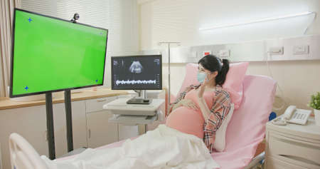 telemedicine concept - asian pregnant woman wearing protective face mask to prevent COVID19 lying in hospital ward has video call with green computer screenの写真素材