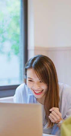 asian young woman using laptop computer makes a deal or wins games with fist gesture by window at homeの写真素材