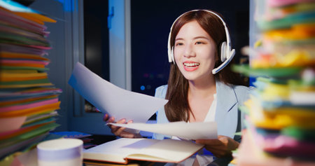Asian overtime businesswoman wearing headset uses laptop computer to join video meeting in office at...