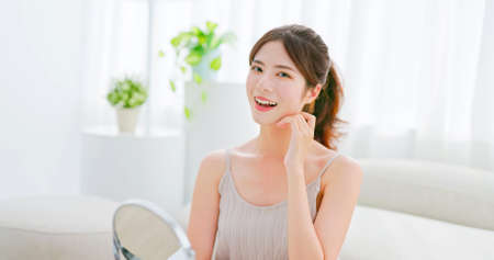 portrait beauty skin care asian woman with brunette ponytail touches her face smiling and looking at you in front of table mirrowの写真素材