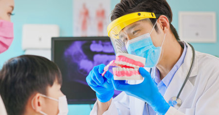 asian dentist with face shield and mask teach boy how to toothbrush brushing teeth use denture model...