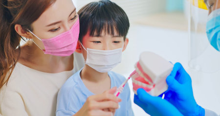 asian dentist with face shield and mask teach boy how to toothbrush brushing teeth use denture model when mother sit by in dental clinic - kid practice itの写真素材