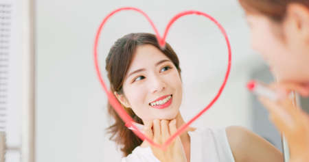 portrait of asian woman with brunette ponytail draws a heart shape use lipstick on the mirror and look at herself feeling confident self loveの写真素材