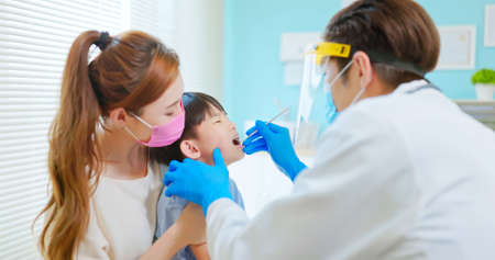 asian dentist with face shield and mask use mouth mirror to exam toothache boy when mother sit by in dental clinic - he looked scaredの写真素材