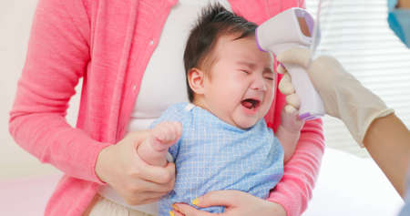 authentic shot of asian mother and infant in pediatrics clinic - doctor examine crying baby health and using thermometer to check body temperature on foreheadの写真素材