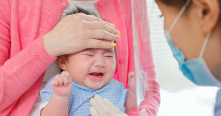 authentic shot of asian mother and infant in pediatrics clinic - doctor examine crying baby health and using thermometer to check body temperature on foreheadの写真素材