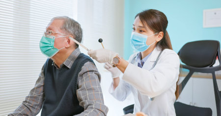 asian female doctor wearing face mask is using tuning fork for hearing tests to elder senior man...