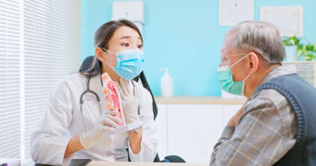 asian female doctor wearing face mask is showing ENT model and explaining to elder senior man patient in hospitalの写真素材