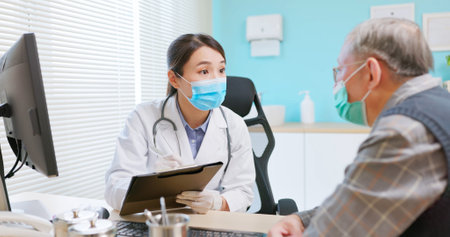 asian female doctor wearing white coat and face mask is diagnosing gray hair elder senior sick male patient in hospital - she takes note on paper documentの写真素材