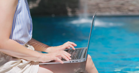 close up of girl is sitting on edge of swimming pool feet underwater in rippled clear blue water and using laptop for workingの写真素材