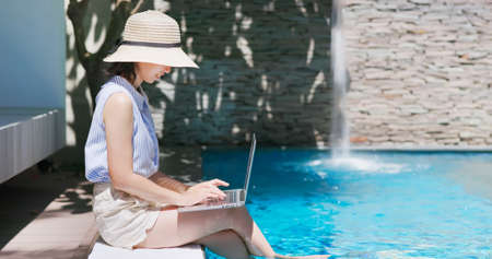 asian girl wearing hat is sitting on edge of swimming pool feet underwater in rippled clear blue water and using laptop for workingの写真素材