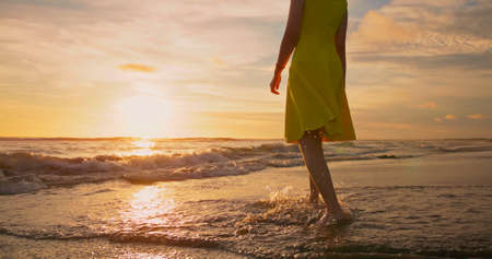close up of woman in beautiful yellow dress feet walking barefoot to beach at golden sunset - Female tourist on summer vacationの写真素材