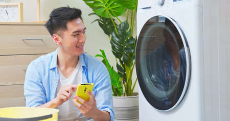 asian man is using phone to control the washing machine when cleaning clothesの写真素材