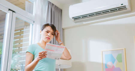 asian woman stand by air conditioner shocked and upset about electricity bill in hand at home - economic inflation conceptの写真素材