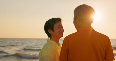 close up of asian elderly carefree couple standing on the beach and smiling at you happily in sunset...