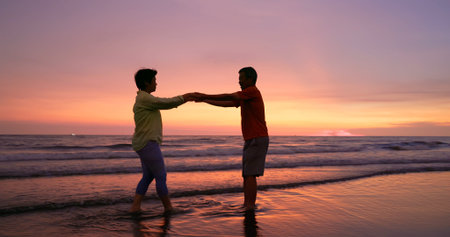 close up of asian elderly carefree couple standing on the beach and smiling at you happily in sunset...