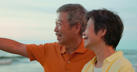 close up of asian elderly carefree couple standing on the beach and smiling at you happily in sunset...