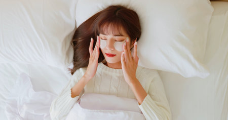happy asian beauty skin care woman gently touching under eye mask on her face satisfied looking in mirror at home during winterの写真素材