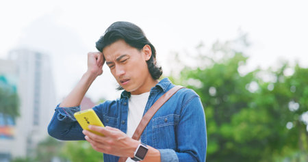asian man walking on the street feeling upset using his smartphone