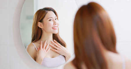 asian cheerful confident woman is looking in mirror and touching her good skin on neckの写真素材