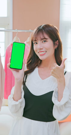 beautiful asian girl selling fashionable clothes on live streamg and she show green screen on smart phoneの写真素材