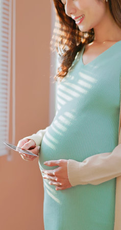 Closeup of Asian pregnant woman standing by the window happily looking at ultrasound photo enjoying her motherhood momentの写真素材