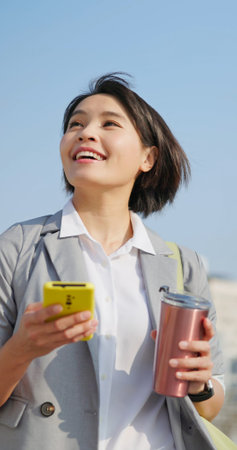 close up asian happy business woman using smartphone and eco cup while walking and commuting in city