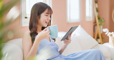 asian woman watching tablet relaxingly drinking tea or coffee sitting on a comfortable sofa