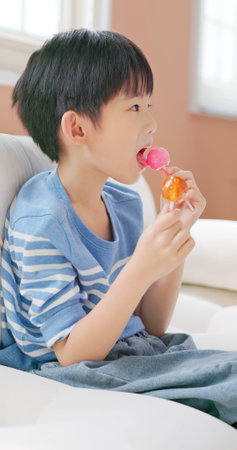 closeup asian children eat unhealthy high calories sugary food sweet candy lollipops sit on sofa which is not good for growing upの写真素材